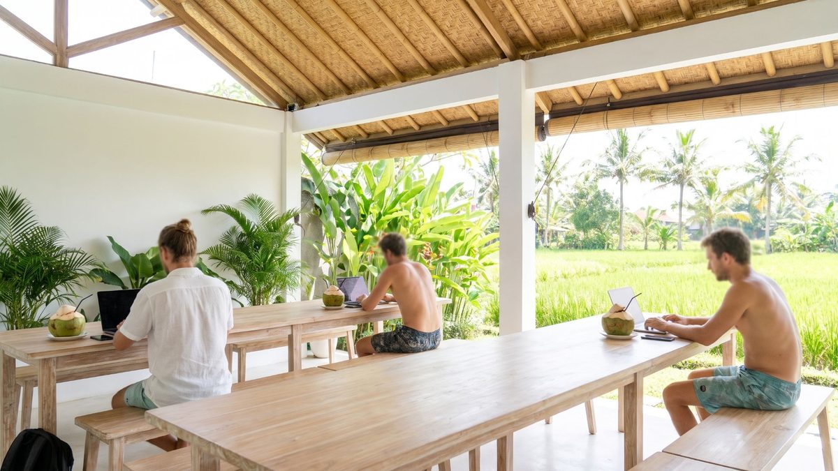 Offener Coworking-Raum unter einem Bambusdach auf Bali: drei Leute arbeiten an Laptops an Holztischen, vor ihnen Trinkkokosnüsse, draußen Palmen und Reisfelder.