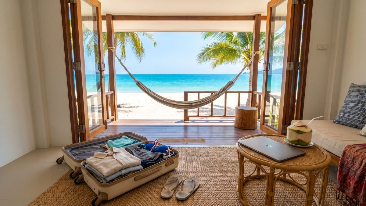 Blick aus einem Strandbungalow durch offene Holztüren auf eine Hängematte zwischen Palmen, weißen Sand und türkises Meer, im Zimmer ein offener Koffer und ein Laptop auf einem Rattantisch.